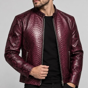 Può includere: Una giacca bordeaux intenso con motivo testurizzato a pelle di serpente, colletto alla coreana e cerniera frontale color argento. Il capo presenta due tasche laterali con chiusura a zip e maniche lunghe, indossato sopra una maglia nera.
