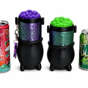 May include: Two black plastic cauldron-shaped containers with iridescent bands labeled "Witches Brew" and removable lids topped with textured purple and green bubbles, positioned between a red Alani Nu energy drink and a green Mountain Dew soda can.
