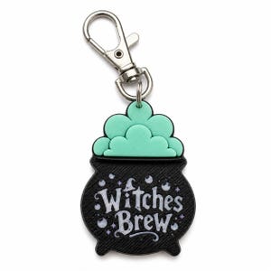 May include: A black cauldron-shaped keychain with a silver-toned metal lobster clasp. The cauldron features a textured surface, a mint green bubbling liquid top, and the white text Witches Brew surrounded by small white and purple star accents.