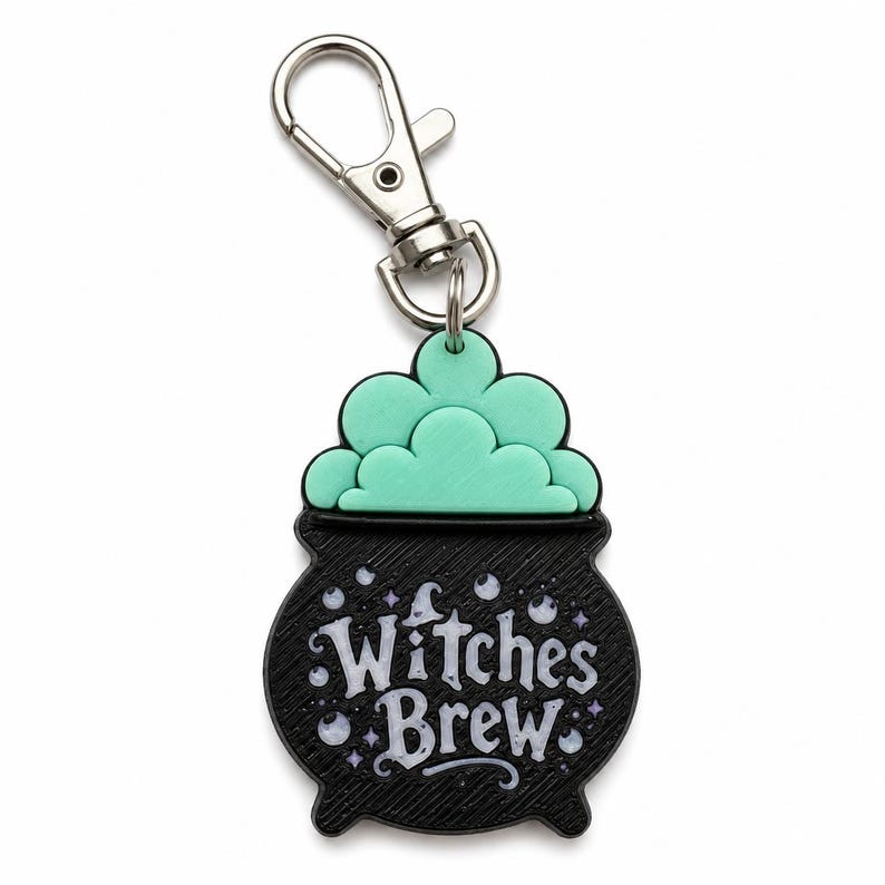 May include: A black cauldron-shaped keychain with a silver-toned metal lobster clasp. The cauldron features a textured surface, a mint green bubbling liquid top, and the white text Witches Brew surrounded by small white and purple star accents.