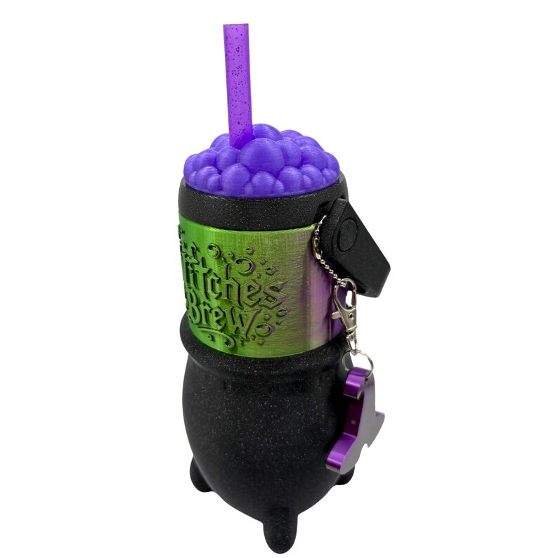 May include: A black cauldron-shaped cup with a textured finish, featuring a green band embossed with the text Witches Brew. The top has purple bubbles and a glittery purple straw. A black handle and a purple metal charm are attached to the side.