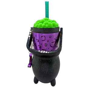 May include: A black cauldron-shaped tumbler with a glittery finish, featuring a purple middle band with embossed bubble details. The top has a bright green textured lid resembling bubbling liquid and a matching teal straw. A black handle is attached.
