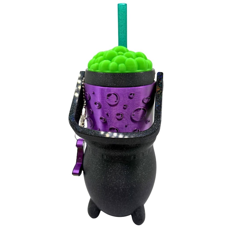 May include: A black cauldron-shaped tumbler with a glittery finish, featuring a purple middle band with embossed bubble details. The top has a bright green textured lid resembling bubbling liquid and a matching teal straw. A black handle is attached.