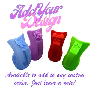 Op de afbeelding: Vier kleurrijke, decoratieve ringhouders in blauw, paars, rood en groen. Elke houder heeft een uniek ontwerp: een bloem, een roos, een strik en een ster. De tekst "Add Your Design" staat erboven.