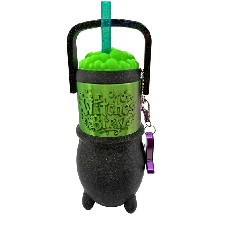 May include: A black cauldron-shaped tumbler with a glittery finish, featuring a metallic green center band embossed with the text Witches Brew. It includes a bright green bubble-textured lid, a translucent green straw, a black handle, and a purple charm.