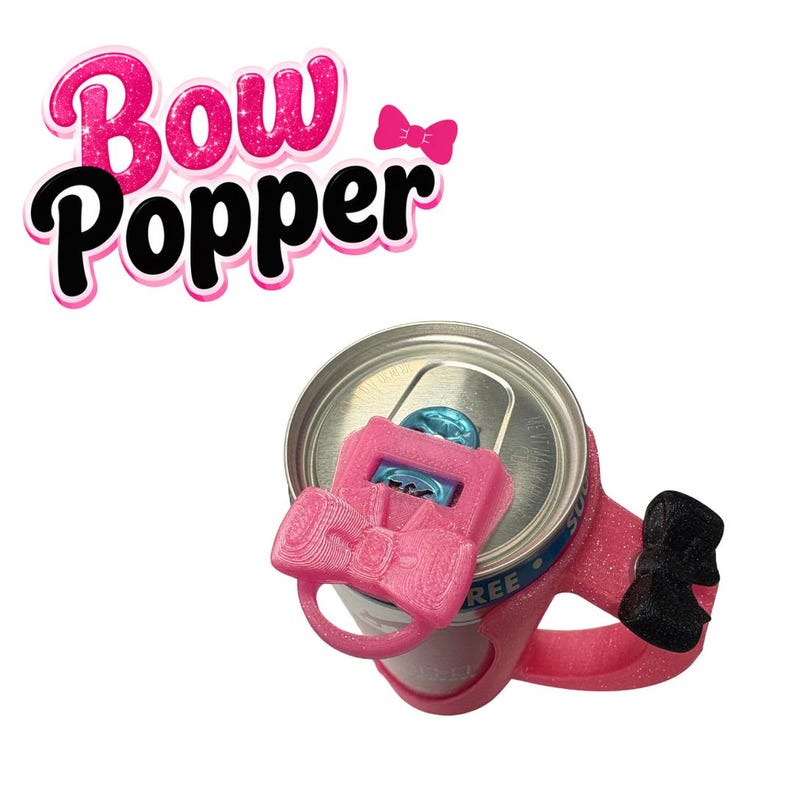 May include: A pink Bow Popper can opener attached to a soda can. The Bow Popper is shaped like a bow and has a handle for easy use. The text "Bow Popper" is in pink with a black outline. The can is silver with blue and white accents.