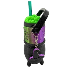 May include: A black cauldron-shaped tumbler with a glittery finish, featuring a metallic green and purple band with text, a bright green bubble-textured lid, a black handle, a green straw, and a small purple witch hat charm on a silver chain.