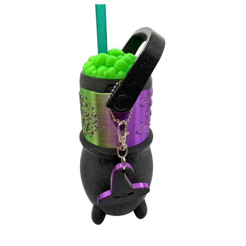 May include: A black cauldron-shaped tumbler with a glittery finish, featuring a metallic green and purple band with text, a bright green bubble-textured lid, a black handle, a green straw, and a small purple witch hat charm on a silver chain.