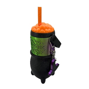 May include: A black cauldron-shaped container with a textured finish, featuring a green metallic band embossed with the text Witches Brew. The top is filled with orange bubbles and an orange straw, with a purple hat charm attached by a chain.