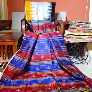 Puede incluir: Un textil colorido con un patrón de rayas en rojo, azul, amarillo y negro. La tela está drapeada sobre una silla de madera. El textil tiene un diseño geométrico y es probablemente una prenda tradicional o una pieza decorativa.