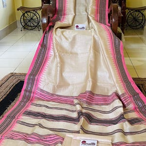 Op de afbeelding: Een beige sari met een opvallende roze en zwarte rand met een geometrisch driehoekspatroon. De stof heeft een gladde, lichtreflecterende afwerking. Een klein wit label met de tekst SPARSH is bevestigd aan de stof nabij het midden.