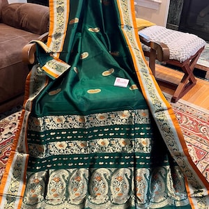 May include: Emerald green sari with gold and orange accents. The fabric features a detailed floral and paisley pattern along the border and lower section. The sari is draped over a chair, showcasing its intricate design and rich color.