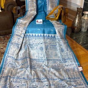 May include: A teal and white patterned textile with intricate designs. The fabric features a detailed pattern of floral and geometric shapes in white against a teal background. The textile is folded and draped, showcasing its texture and design. A yellow fabric is folded on top.