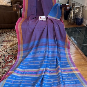 Pu&ograve; includere: Un tessuto vibrante con una tavolozza di colori viola, blu, rosa e giallo. Il tessuto &egrave; drappeggiato su una sedia, mostrando un motivo a righe con dettagli a diamante. I bordi sono rifiniti con nappe. Il tessuto ha il marchio "SPARSH" su un'etichetta.