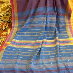 Pu&ograve; includere: Un tessuto intrecciato nei toni del blu, viola e rosa. Il tessuto presenta strisce orizzontali e motivi geometrici. I bordi sono rifiniti con nappe. Il tessuto &egrave; probabilmente uno scialle o un copriletto decorativo.