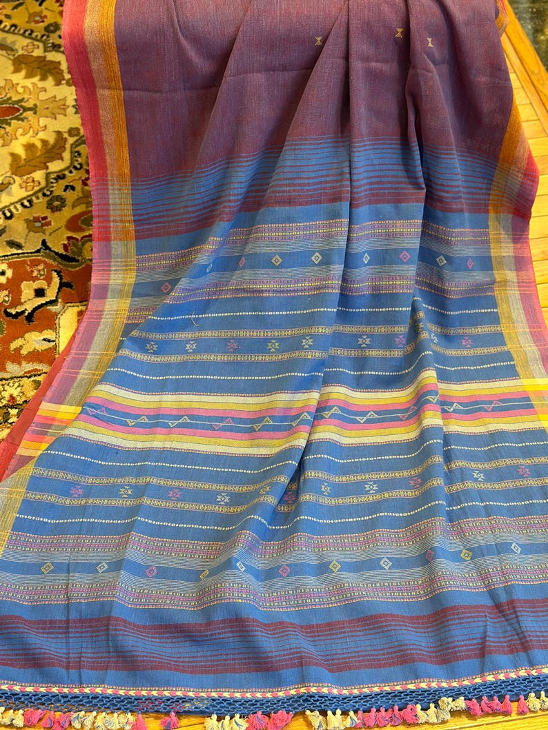 Pu&ograve; includere: Un tessuto intrecciato nei toni del blu, viola e rosa. Il tessuto presenta strisce orizzontali e motivi geometrici. I bordi sono rifiniti con nappe. Il tessuto &egrave; probabilmente uno scialle o un copriletto decorativo.