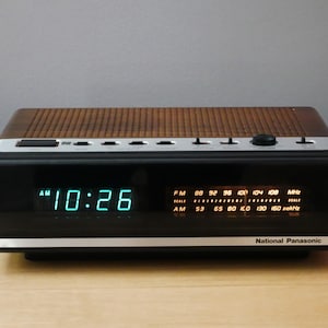 Op de afbeelding: Een vintage National Panasonic digitale klokradio. De klok geeft "10:26" weer in blauwe cijfers. De radio heeft een bruine, geperforeerde bovenkant, een zwart voorpaneel en zilveren accenten. Het toont ook FM- en AM-radiofrequenties.
