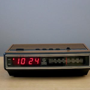 May include: A vintage General Electric digital alarm clock. The clock has a black base with a wood-grain top and a red LED display showing the time as 10:24. The clock also has an AM/FM radio dial.