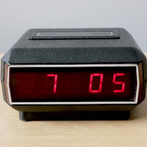 May include: A black digital alarm clock displaying the time 7:05 in red numerals. The clock has a rectangular shape with a silver trim around the display. The clock is sitting on a light-colored surface.