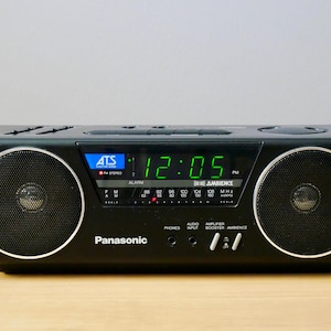 Puede incluir: Radio despertador estéreo AM/FM Panasonic negro con una pantalla digital que muestra las 12:05 PM. La radio tiene dos altavoces circulares y varios botones y entradas. El texto "THE AMBIENCE" es visible.
