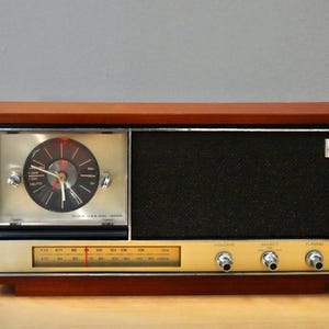 Puede incluir: Una radio rectangular de madera con un reloj analógico integrado en el lado izquierdo y una rejilla de altavoz negra a la derecha. El panel frontal presenta una pantalla de radiofrecuencia dorada, tres perillas de control plateadas y el texto Clock Radio Japan.