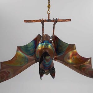 May include: A colorful metal bat hanging from a ceiling with a brown wooden dowel. The bat has iridescent wings with shades of blue, green, orange, and brown.