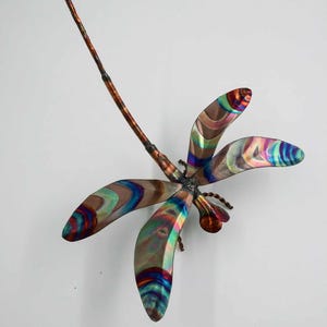 May include: A colorful metal dragonfly wall sculpture with iridescent wings. The dragonfly is made of copper and has a long, thin body.