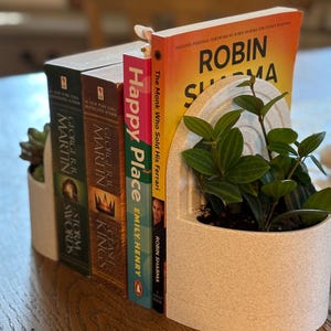 Op de afbeelding: Een decoratieve boekensteun met een ingebouwde plantenbak, met verschillende boeken met kleurrijke ruggen. De boeken bevatten titels als "Happy Place" en "The Monk Who Sold His Ferrari". De plantenbak bevat groene planten.