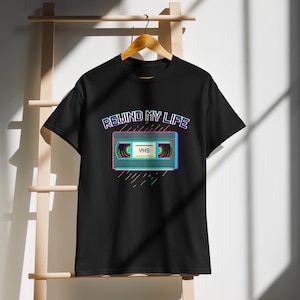 Puede incluir: Camiseta negra con un diseño retro de cinta VHS y el texto "REWIND MY LIFE" en fuente de neón. La cinta VHS tiene la palabra "VHS". El diseño utiliza colores azul, rosa y amarillo.