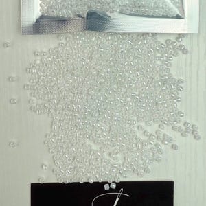 May include: A close-up image of a bag of white seed beads. The beads are small and round, and they are scattered on a white surface.