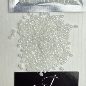 May include: A bag of white seed beads, with a small pile of beads spilled out on a white surface. The bag has a label that says "Made Whitlock Timeless Luxury".
