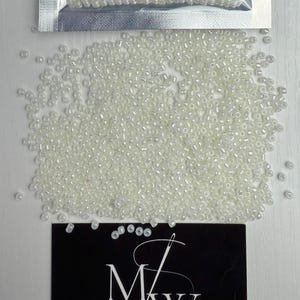 Puede incluir: Una imagen de primer plano de una gran cantidad de pequeñas cuentas redondas blancas. Las cuentas están esparcidas sobre una superficie blanca. Las cuentas están en una bolsa de plástico transparente con un respaldo plateado.