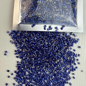 Op de afbeelding: Een close-up afbeelding van een grote hoeveelheid kleine, blauwe glazen kralen. De kralen zijn cilindrisch van vorm en hebben een metallic glans.