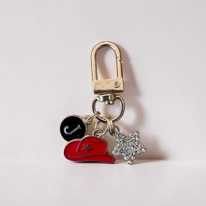 May include: A gold-colored keychain with a lobster clasp, featuring three charms: a red cowboy hat with a star, a black circle with a curved white line, and a rhinestone starburst.