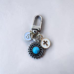 May include: Silver keychain with a lobster clasp, featuring charms. Charms include a turquoise stone pendant, a white circle with a black cross, and a white circle with the letter "S".