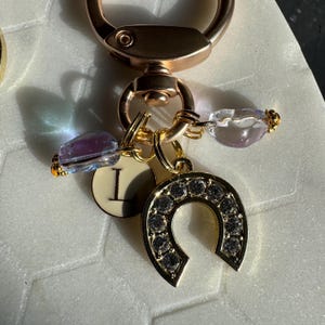 May include: A gold-colored keychain with a horseshoe charm encrusted with small, clear crystals. The keychain also features a charm with the letter "I" and two decorative beads in shades of purple and clear. The clasp is a rose gold color.