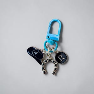 May include: A blue keychain with a blue clasp, featuring charms of a navy cowboy hat with a star, a horseshoe, and a black circle with the letter "R". The horseshoe charm is silver with black accents.