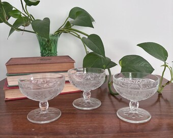 May include: Three clear glass dessert bowls featuring a delicate embossed floral pattern on the exterior. Each bowl is supported by a short, sturdy pedestal base with a circular foot, showcasing a consistent design across all three pieces.