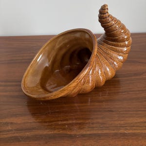 May include: A brown ceramic cornucopia, also known as a horn of plenty, with a glossy finish. The horn curves upward and has detailed ridges and a wide opening. The interior is a lighter shade of brown.