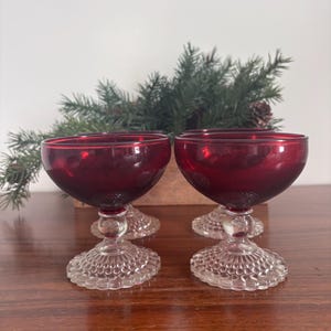 May include: Four ruby red stemmed glasses with a clear, textured base. The glasses have a rounded bowl shape. The background features a wooden box with evergreen branches and a pinecone.