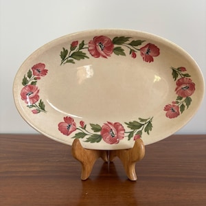 May include: Oval serving platter with a cream-colored base, adorned with a floral pattern of red poppies and green leaves. The platter is displayed on a wooden stand, showcasing its decorative design.
