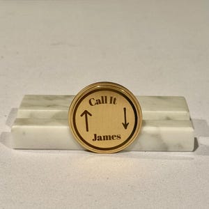May include: A gold-colored round compass with the text "Call It" and "James" engraved on it. The compass has two arrows pointing up and down.