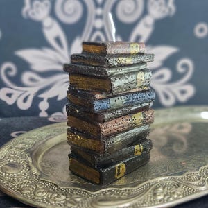 Pode incluir: Uma vela em forma de pilha de livros. A vela, com um pavio branco, é projetada para se assemelhar a uma coleção de livros antigos em várias cores, incluindo marrom, dourado e azul. Está colocada numa bandeja prateada com um design ornamentado.