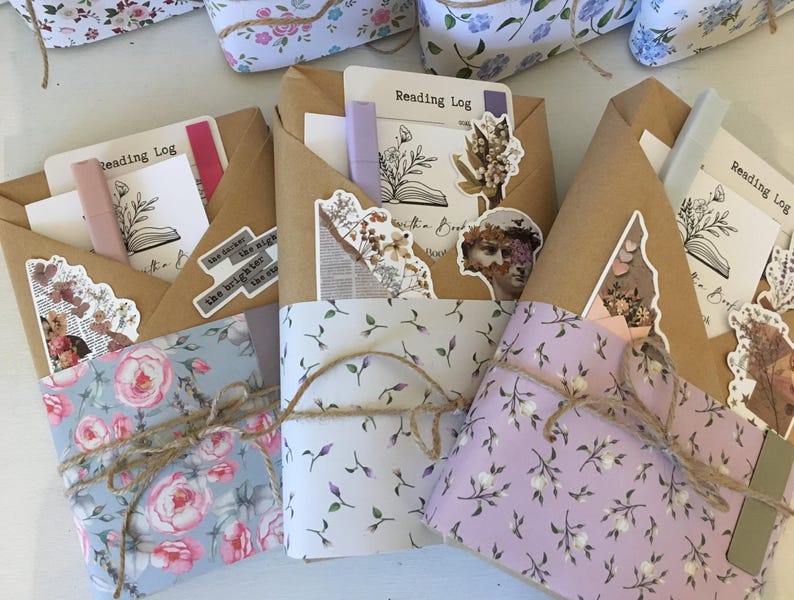 May include: Three brown paper wrapped journals with floral patterned paper on the front. Each journal has a sticker with the text "Reading Log" and a small butterfly graphic. The journals are tied with twine.
