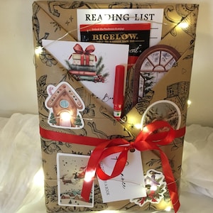 May include: A wrapped gift with a brown paper cover, adorned with a red ribbon and fairy lights. The package includes a "Reading List" and a Bigelow tea packet. Decorative stickers and a red pen are visible, suggesting a book-themed gift.