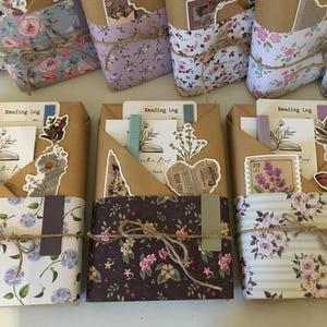 May include: Three brown paper-wrapped journals with floral patterned covers. Each journal has a pocket on the front with a variety of decorative elements, including paper clips, stickers, and a small pen. The journals are labeled "Reading Log".