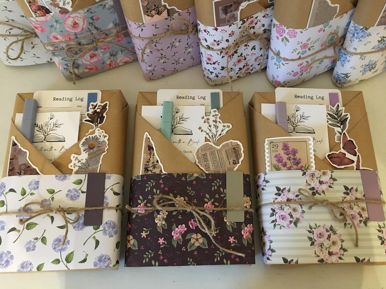 May include: Three brown paper-wrapped journals with floral patterned covers. Each journal has a pocket on the front with a variety of decorative elements, including paper clips, stickers, and a small pen. The journals are labeled "Reading Log".