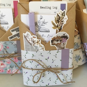 May include: Three brown paper-wrapped journals with floral patterned paper covers. Each journal has a different floral pattern and a handwritten title "Reading Log" on the front. The journals are tied with twine and decorated with stickers and other embellishments.