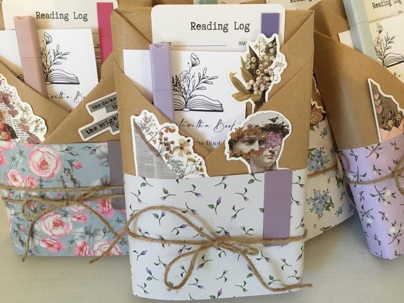 May include: Three brown paper-wrapped journals with floral patterned paper covers. Each journal has a different floral pattern and a handwritten title "Reading Log" on the front. The journals are tied with twine and decorated with stickers and other embellishments.