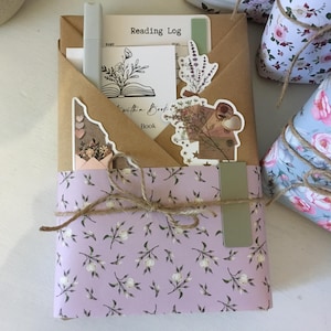 May include: A book wrapped in a light purple paper with a floral pattern. The book is tied with twine and has a brown paper envelope on top with a sticker that says "Reading Log".